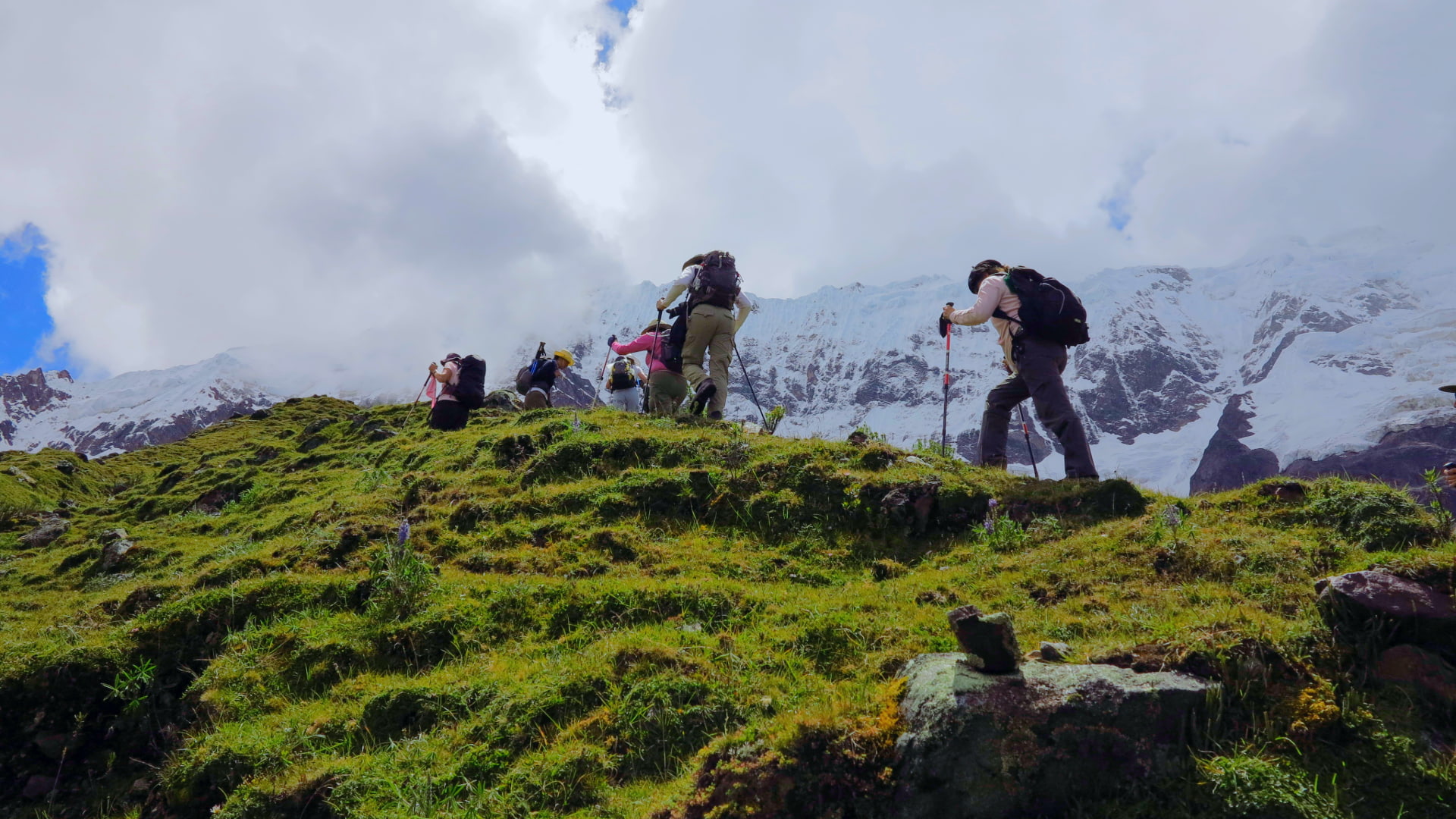 Hikers climbing a steep grassy hill on the Salkantay Trek, surrounded by vibrant greenery and snow-capped peaks in the background under a cloudy sky.