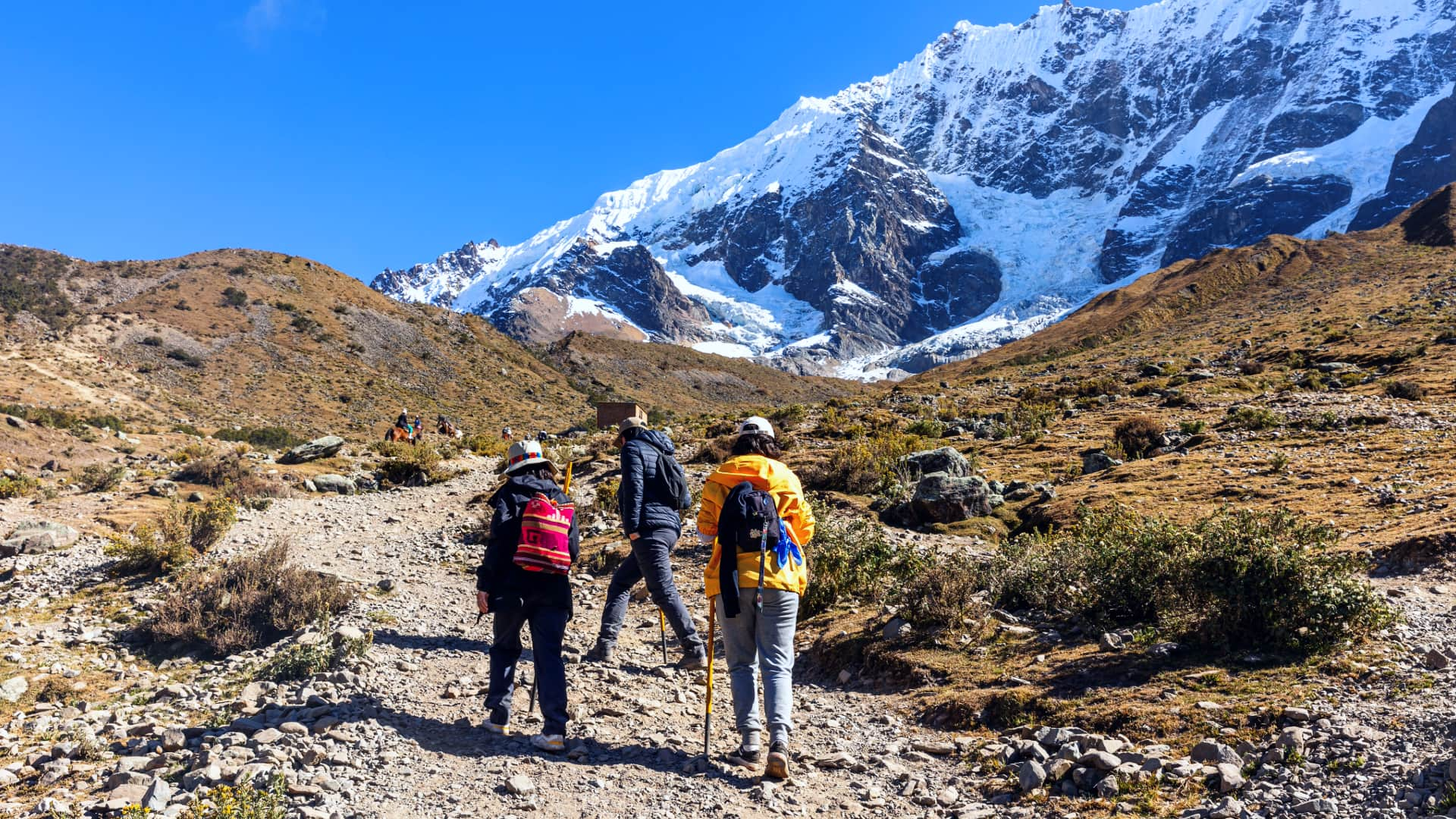 Hikers walking along the rocky trail of the Salkantay Trek, with towering snow-capped peaks of the Peruvian Andes in the background beneath a clear sky.