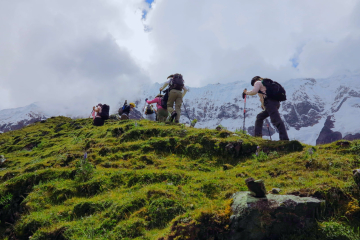 Hikers climbing a steep grassy hill on the Salkantay Trek, surrounded by vibrant greenery and snow-capped peaks in the background under a cloudy sky.