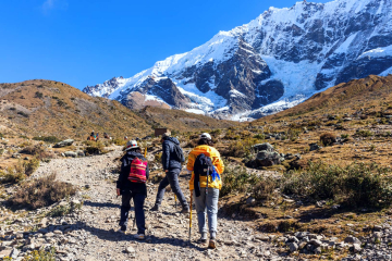 Hikers walking along the rocky trail of the Salkantay Trek, with towering snow-capped peaks of the Peruvian Andes in the background beneath a clear sky.